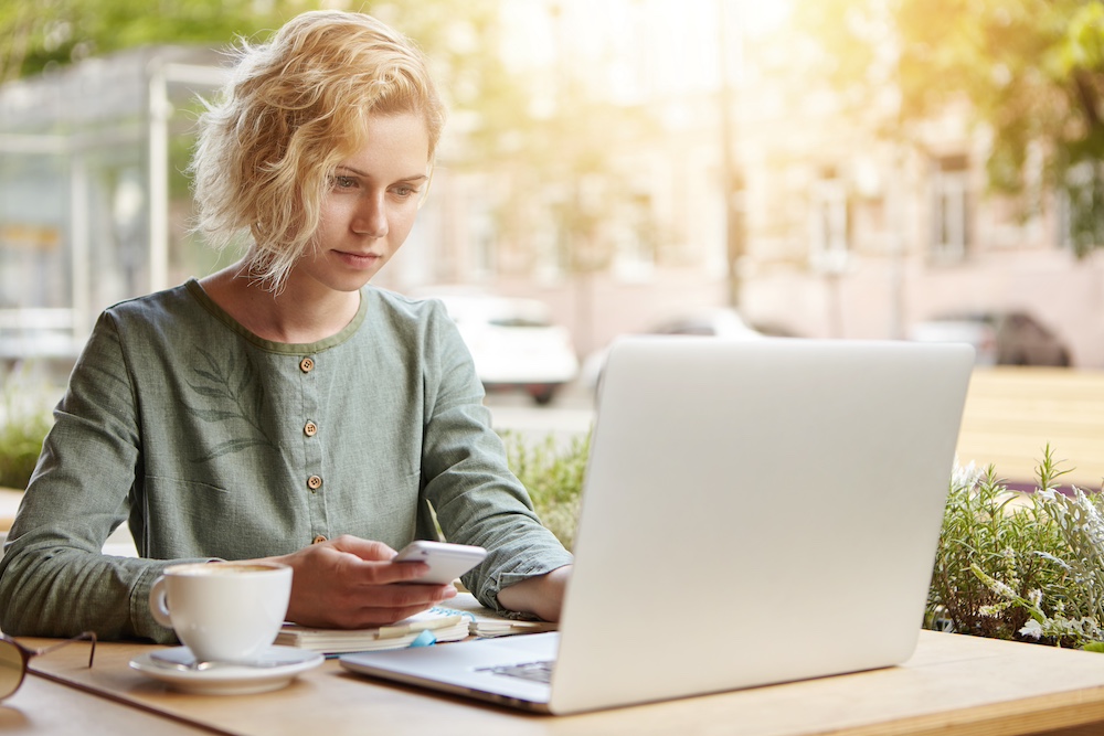 Beautiful female with trendy hairstyle wearing elegant blouse sitting at outdoor cafe drinking coffee surounded with modern gadgets. Young blonde lady holding cell phone in hand searching information